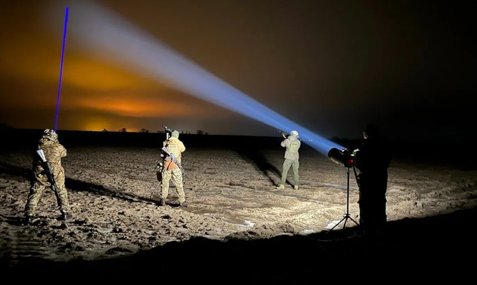 Nighttime military training: four soldiers in a desert, one aiming a blue beam from a spotlight across the ground, with a fourth person operating a large light on a tripod nearby